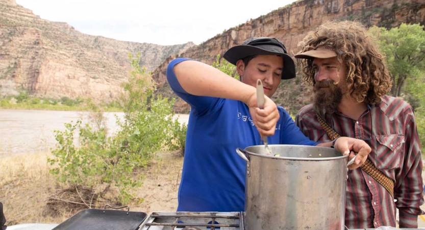 Two people use a large pot to prepare food. Behind them is a river and tall canyon walls. 